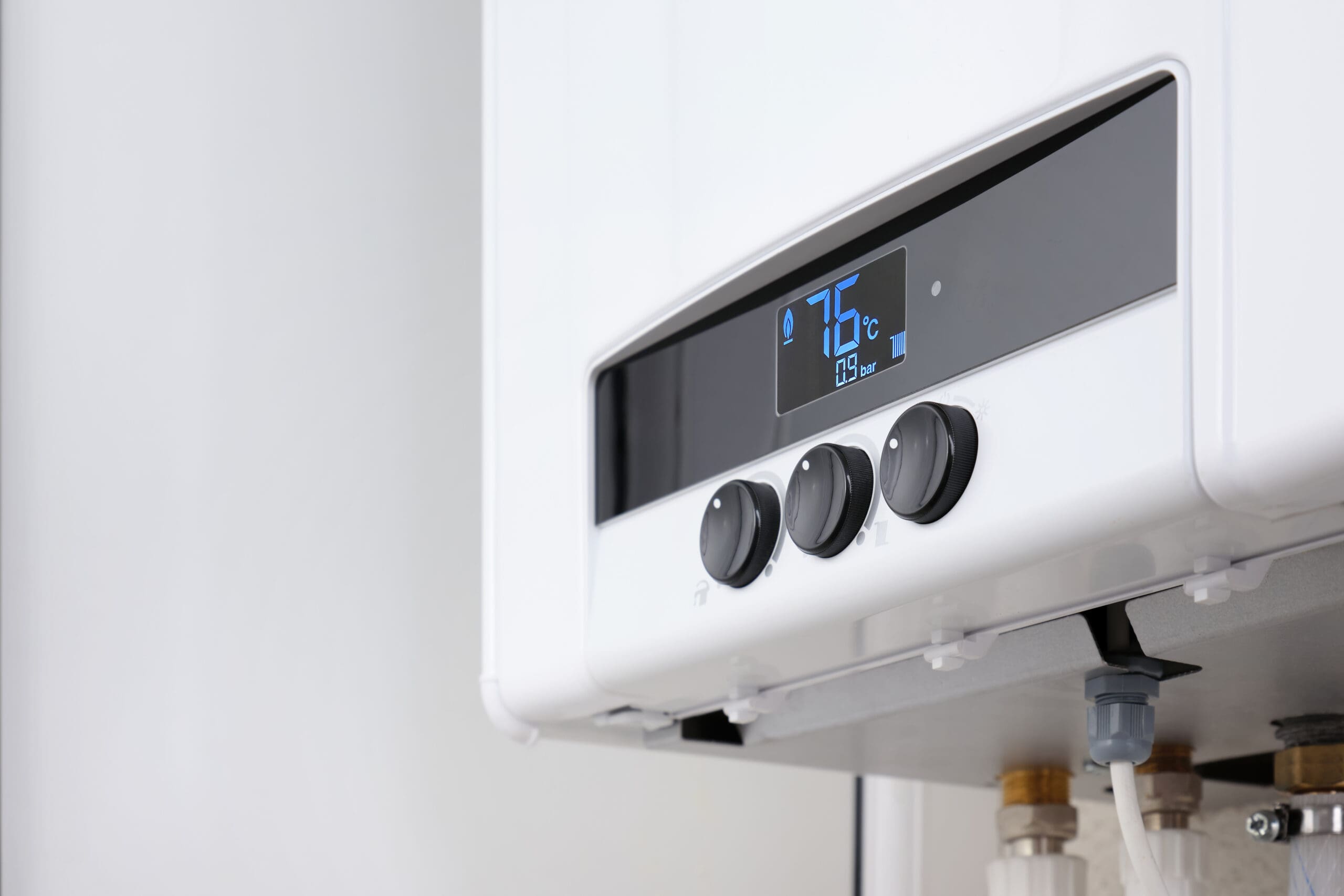 Close-up of a white modern gas boiler with three black control knobs and a digital display showing the temperature at 75°C. Pipes and fittings are partially visible underneath the unit.