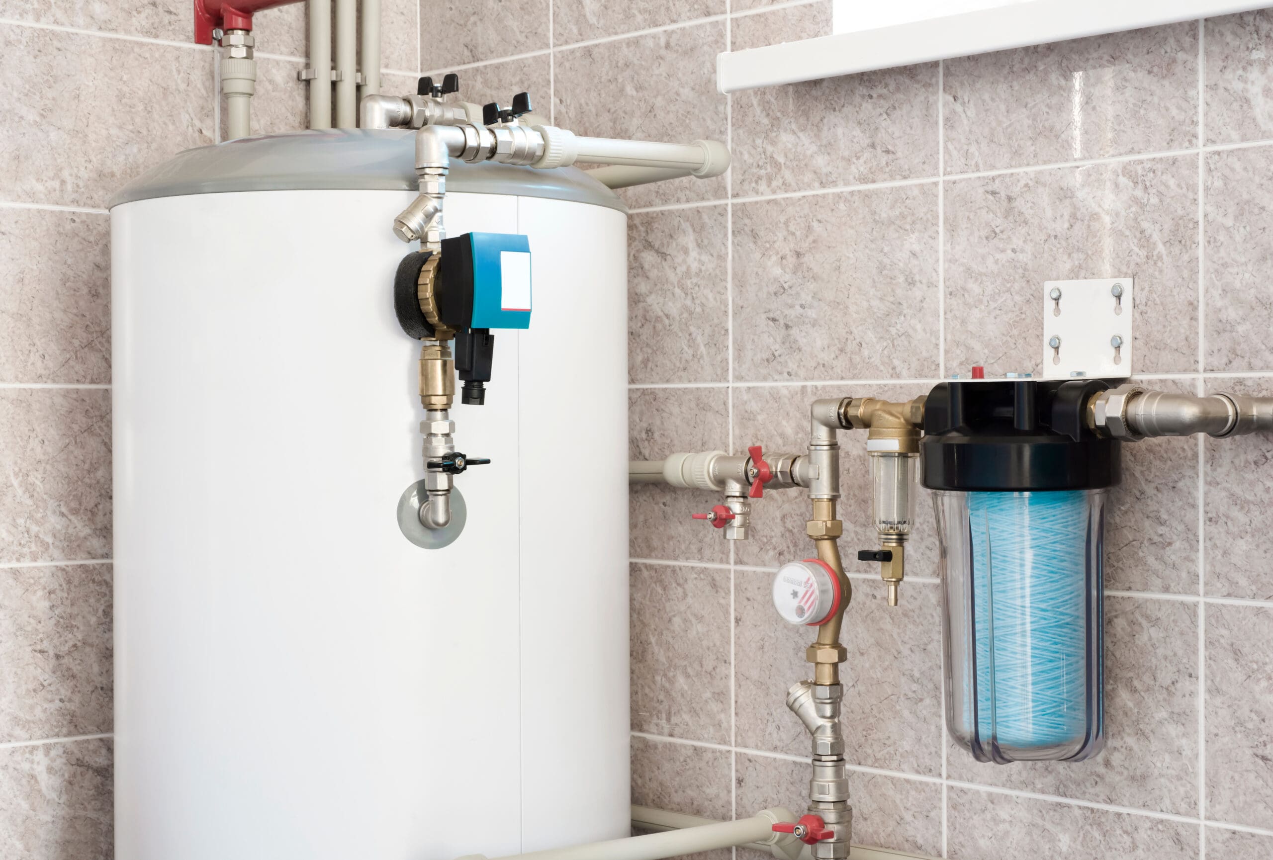 Large white water heater with pipes and valves is mounted on a tiled wall. Next to it, a blue water filter housing is attached to the plumbing. The setup appears clean and organized.