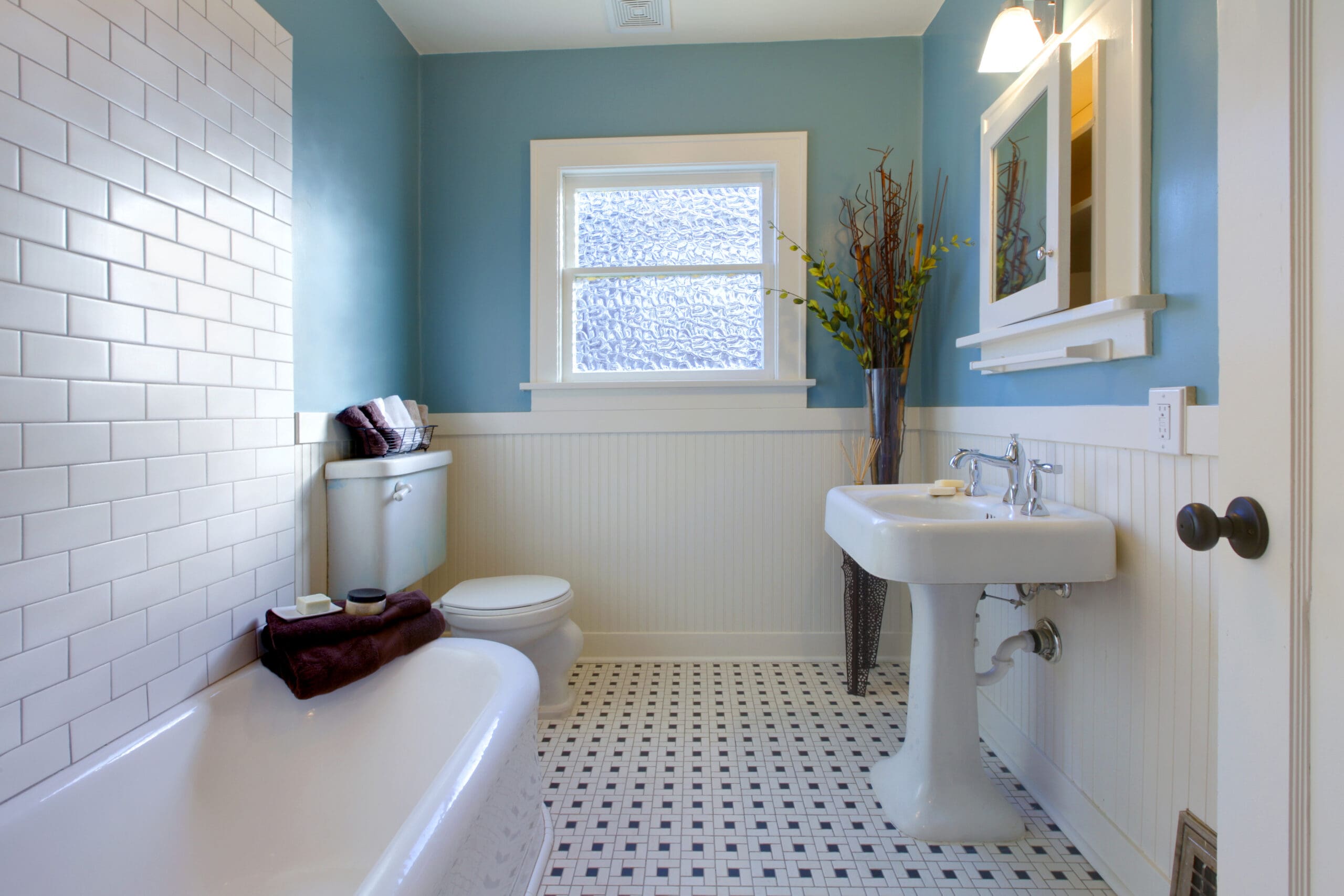 Bright, clean bathroom with a white pedestal sink, toilet, bathtub, tiled floor, blue walls, and white wainscoting. A window lets in light, and a vase with tall branches decorates the corner.