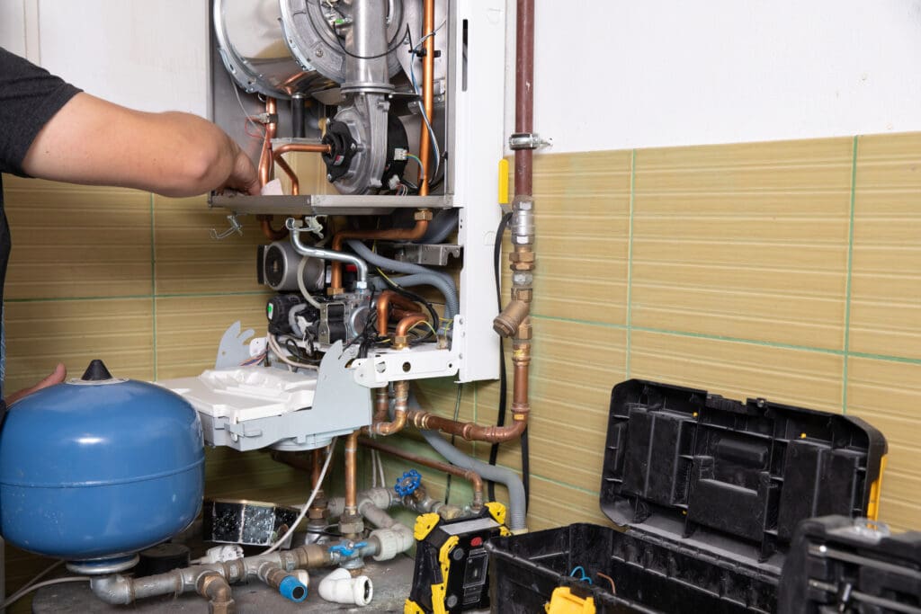 A person is repairing a gas boiler mounted on a tiled wall, with the boiler cover open showing internal parts. A blue expansion tank and an open toolbox with tools are visible on the floor nearby.