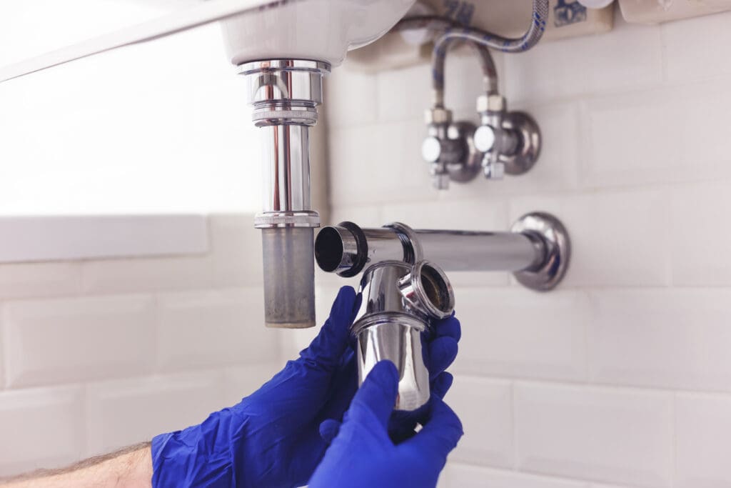 A person wearing blue gloves disassembles a chrome P-trap pipe under a bathroom sink, exposing the drain for cleaning or repair, with white tiled walls in the background.
