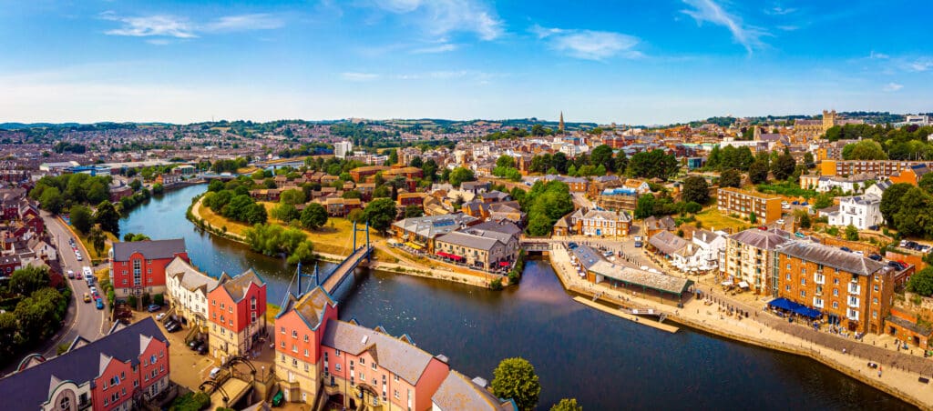 Aerial view of a river winding through a city with colorful buildings, green trees, and a blue sky. People walk along the riverside, and bridges connect both sides of the city.