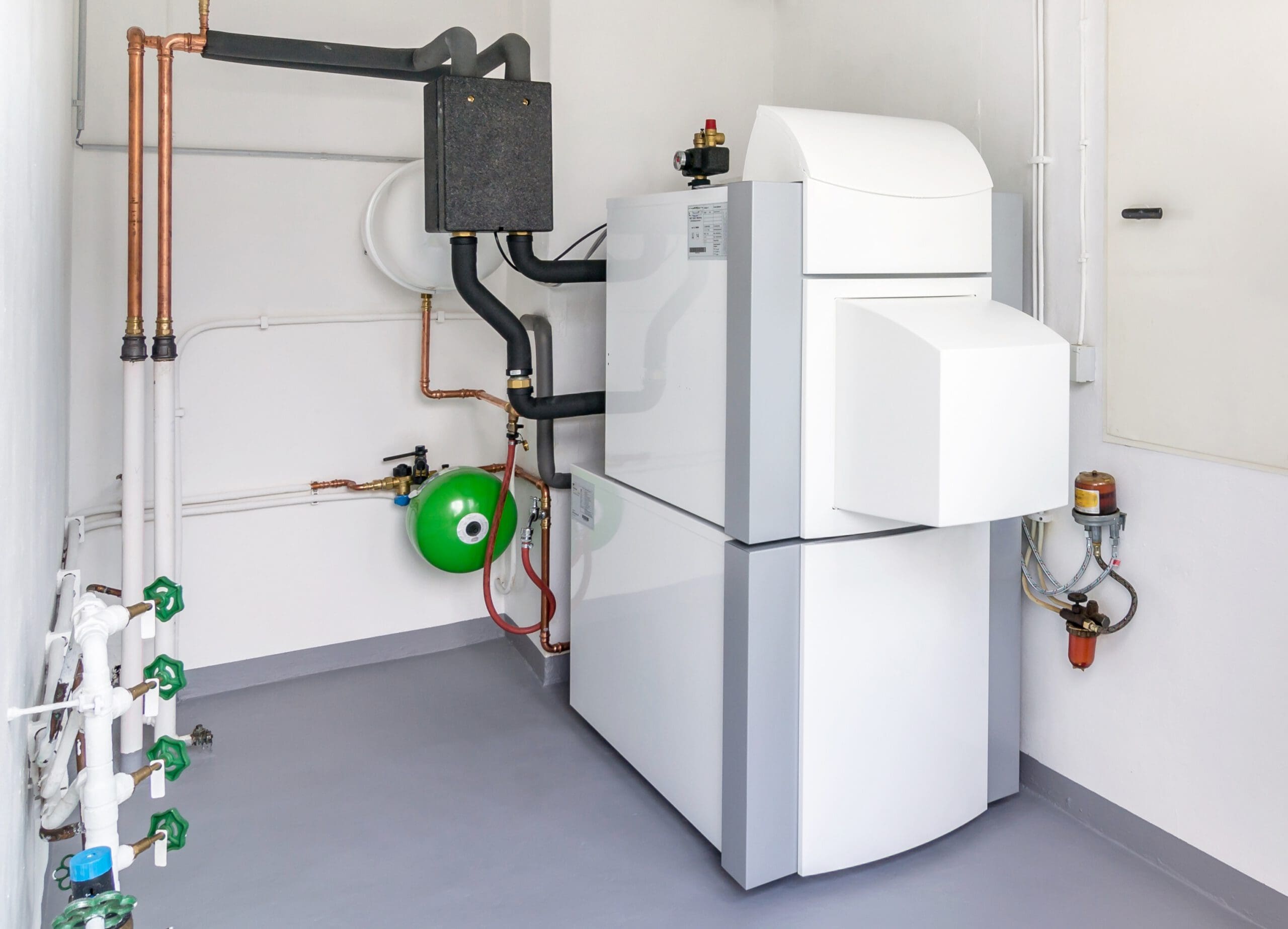 A modern white boiler or heating system with pipes, valves, and a green pressure vessel installed in a clean, white utility room with a gray floor.