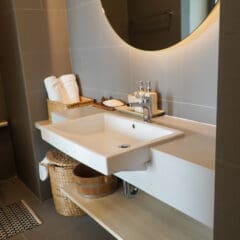 Modern bathroom with a large round mirror above a white rectangular sink, wicker baskets, rolled white towels, and a wooden shelf beneath the countertop. The space is clean and decorated in neutral tones.