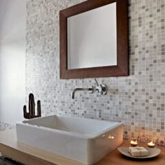 A modern bathroom features a rectangular white sink on a wooden counter, a wall-mounted faucet, a square mirror with a wooden frame, mosaic tile backsplash, and a plate with candles beside the sink.