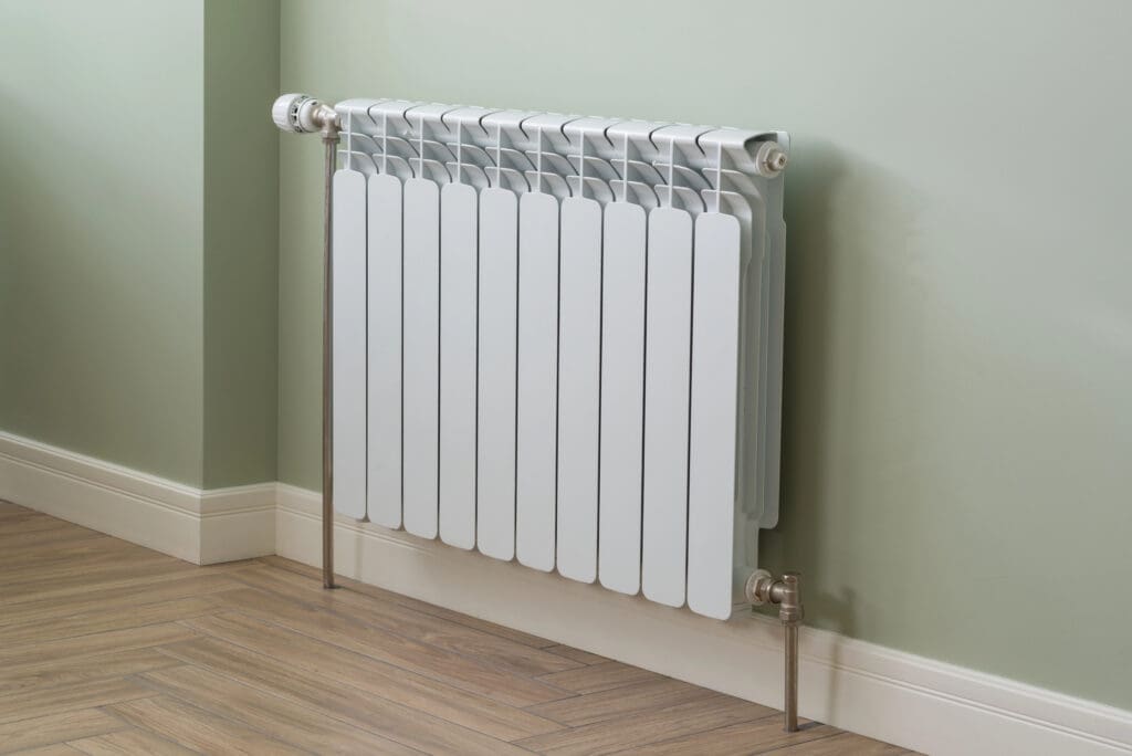 A modern white radiator is mounted on a light green wall above wood-patterned flooring, with metal pipes connected at the top and bottom corners.