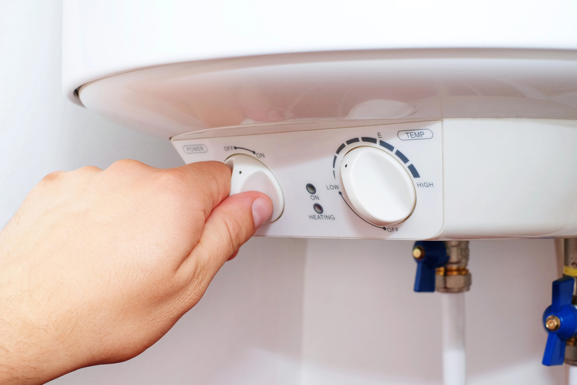 A hand adjusts the control knob on a white water heater, setting it to heating mode. The panel shows power and temperature settings, with blue and yellow valves visible below the unit.