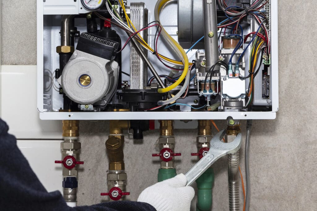 A person wearing a glove uses a wrench to adjust pipes connected to an open wall-mounted boiler, revealing internal components, wires, and valves.