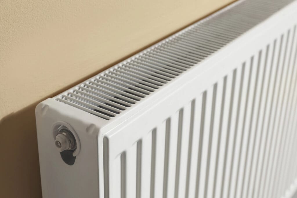 A close-up view of a white metal radiator attached to a cream-colored wall, showing the vented top and side with a valve on the corner.