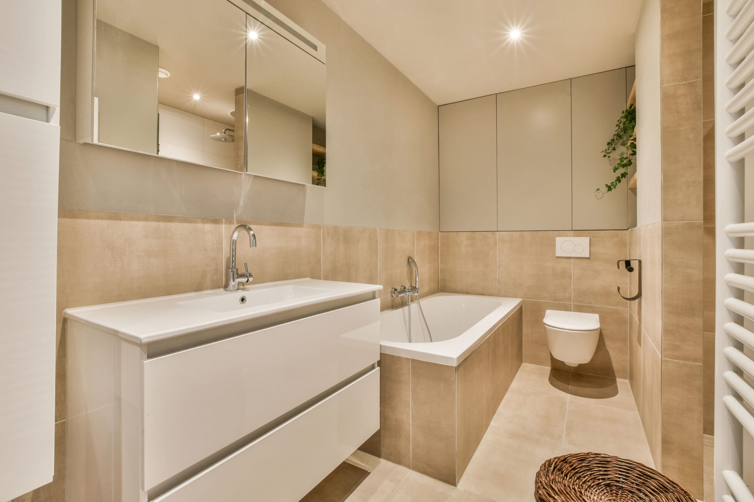 Modern bathroom with beige tiles, white vanity with sink, wall-mounted mirror, built-in bathtub, wall-hung toilet, overhead cabinets, a small plant, and a wicker basket on the floor.