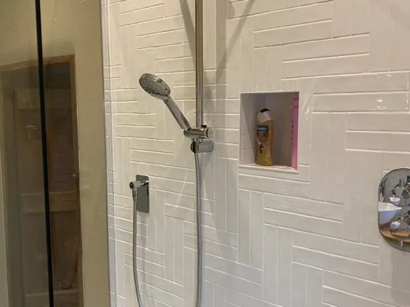 A modern shower with a handheld showerhead, a built-in wall shelf containing a bottle of shampoo and a pink object, white herringbone tiles, and a glass shower door.