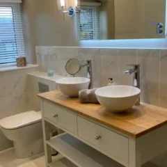 Modern bathroom with a wooden countertop, two white vessel sinks, two chrome faucets, a round mirror, a rolled towel, and a toilet next to a window with white blinds. The space is bright and clean.