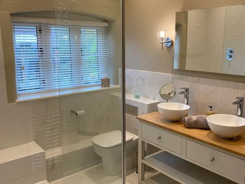 Modern bathroom with a glass shower, white toilet, double sinks on a wooden countertop, large mirror, towels, wall lamp, and a window with white blinds letting in natural light.