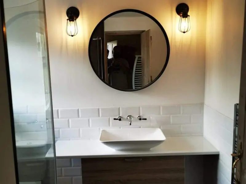 A modern bathroom with a round mirror, two wall-mounted light fixtures, a white rectangular sink, and white subway tile backsplash above a wooden vanity. A shower is visible on the left side.