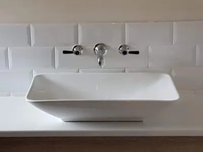 A modern white rectangular vessel sink with two black-handled wall-mounted faucets and a white tiled backsplash.
