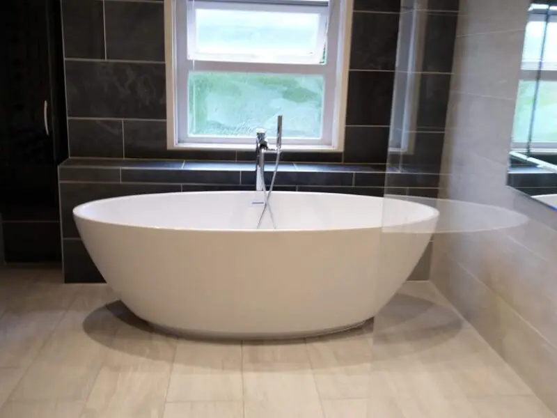 A modern bathroom with a large white freestanding bathtub, light beige floor tiles, dark wall tiles, a window above the tub, and a glass shower partition on the right.