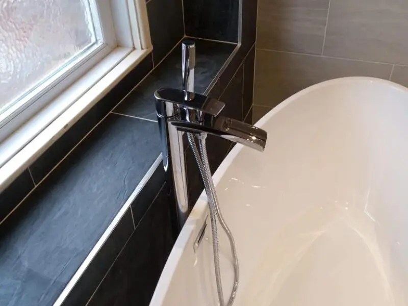 A modern chrome bathtub faucet with a handheld shower attachment is installed next to a white bathtub, near a tiled wall and a frosted window.
