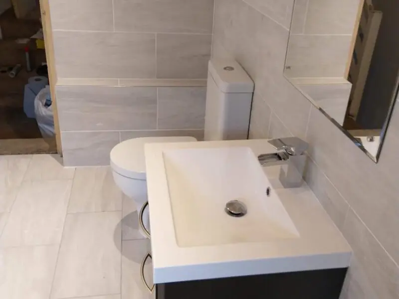 A modern bathroom with light gray tiled walls and floor, featuring a white toilet, a white rectangular sink with a chrome faucet, and a large mirror above the sink.
