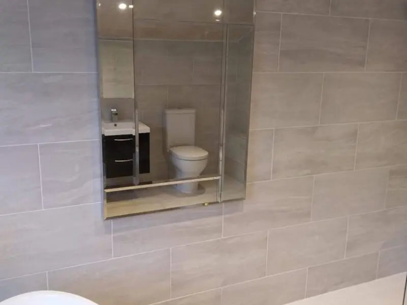 A bathroom with light gray tiled walls. A mirrored cabinet reflects a toilet and a sink with a cabinet beneath, which are not directly visible in the main view.