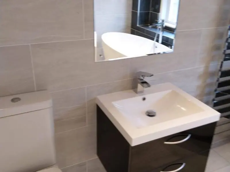 A bathroom with a white toilet, a modern rectangular sink with a cabinet, a wall-mounted faucet, and a mirror reflecting part of a bathtub. The walls are tiled in light gray.