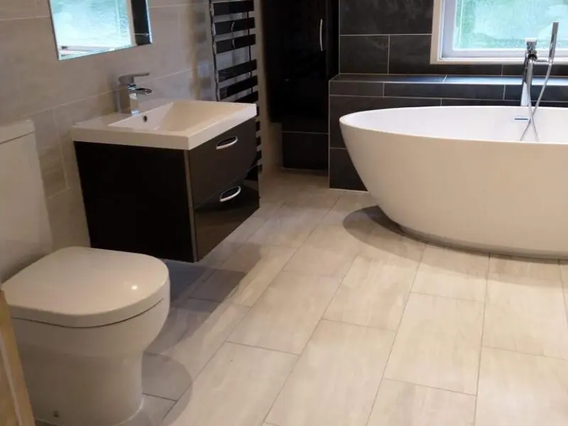 Modern bathroom with light-colored tile floor, a white toilet, floating vanity with sink, black towel radiator, and a large freestanding white bathtub near a window.
