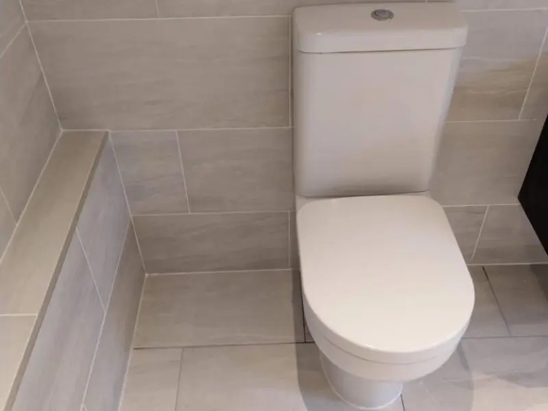 A modern white toilet in a bathroom with light beige tiles on the floor and walls. There is a tiled ledge to the left of the toilet.