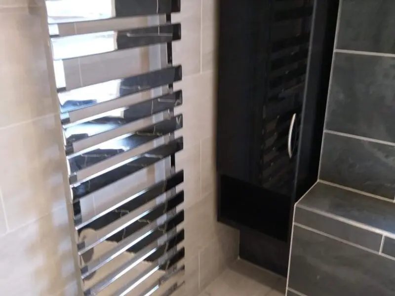 A modern bathroom with a shiny, wall-mounted chrome towel radiator next to dark gray tiled walls and a black cabinet with a silver handle.