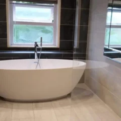 Modern bathroom with a white freestanding bathtub, floor-mounted faucet, dark tile accent wall, light tile floor, a window above the tub, and a large rectangular mirror on the right wall.