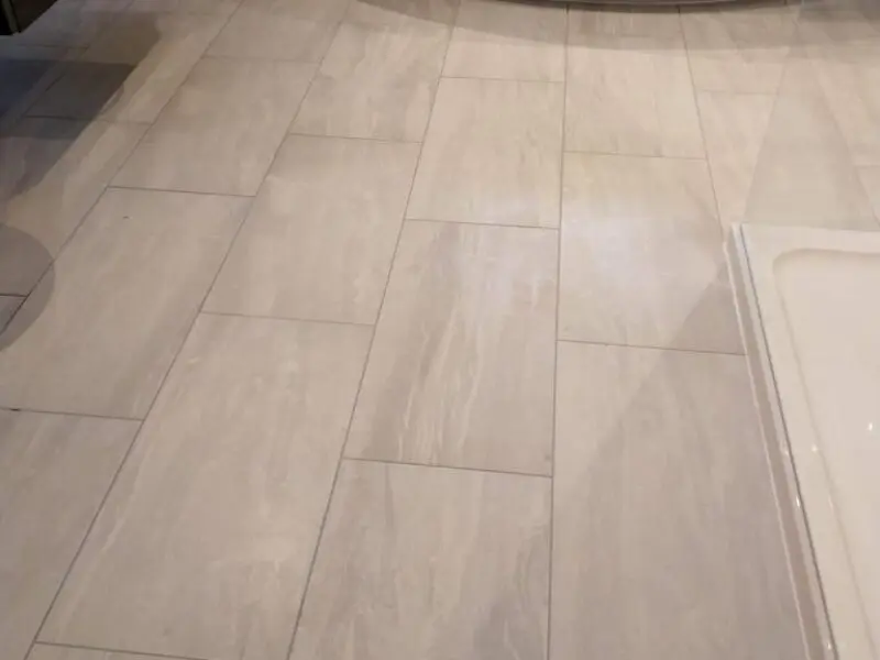 Light-colored rectangular tiles are laid in a staggered pattern on a floor. The image also shows the edge of a white shower tray on the right side. The overall appearance is clean and modern.