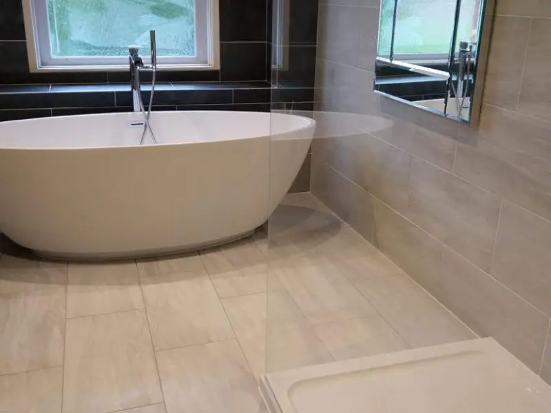 Modern bathroom with a white freestanding bathtub near a window, beige floor and wall tiles, a glass shower partition, and a large mirror above the sink.