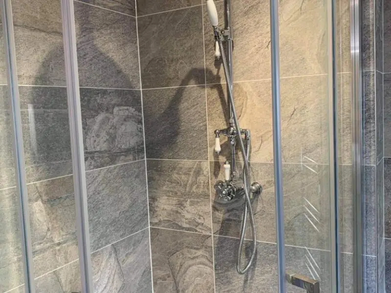 A tiled shower with a glass door, featuring a silver showerhead and handle attached to the wall. The tiles have a gray marble pattern, and light casts a shadow on the wall.