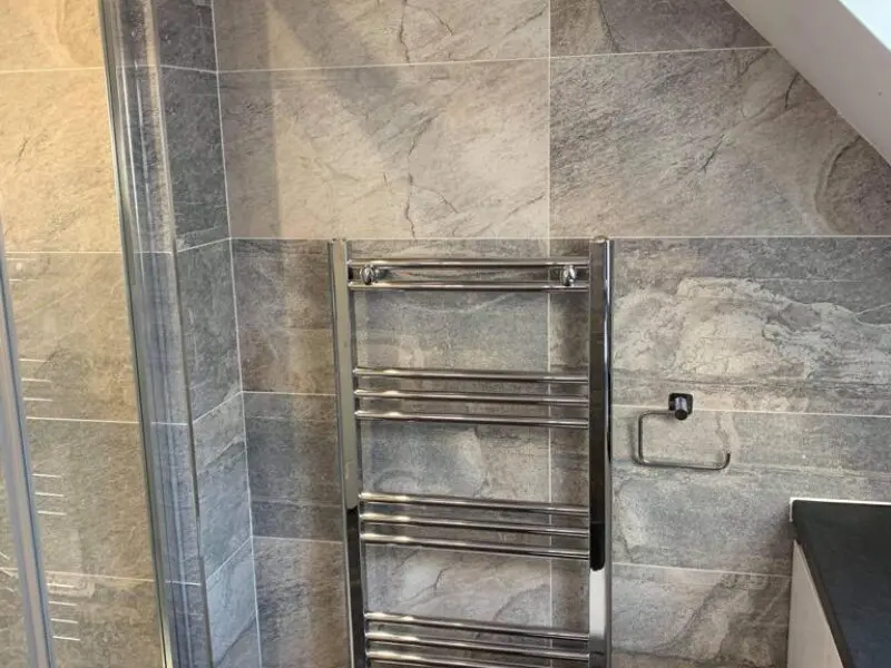 A modern bathroom with gray stone tiles, a chrome towel rack, a toilet paper holder mounted on the wall, and a countertop on the right side.