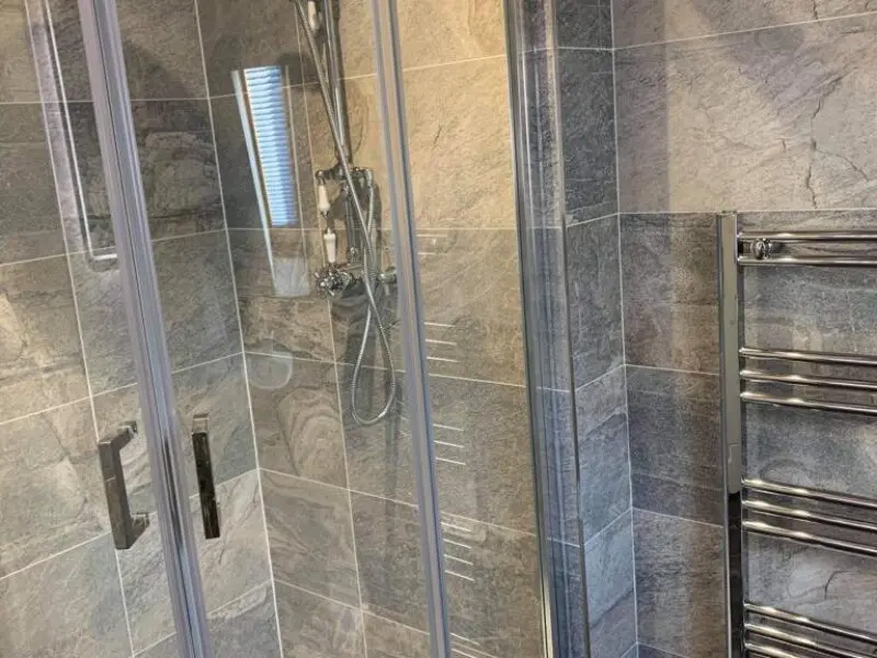 A modern bathroom with gray stone tiles, a glass shower enclosure with metal handles, and a chrome towel radiator on the right side.
