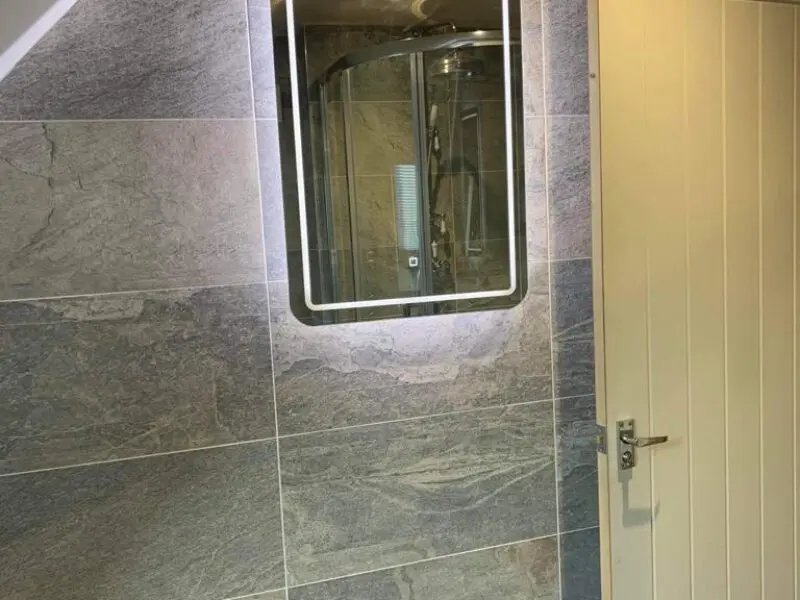 A modern bathroom with gray tiled walls, a rectangular mirror with backlighting, and a light-colored wooden door with a silver handle. The reflection in the mirror shows a glass shower enclosure.