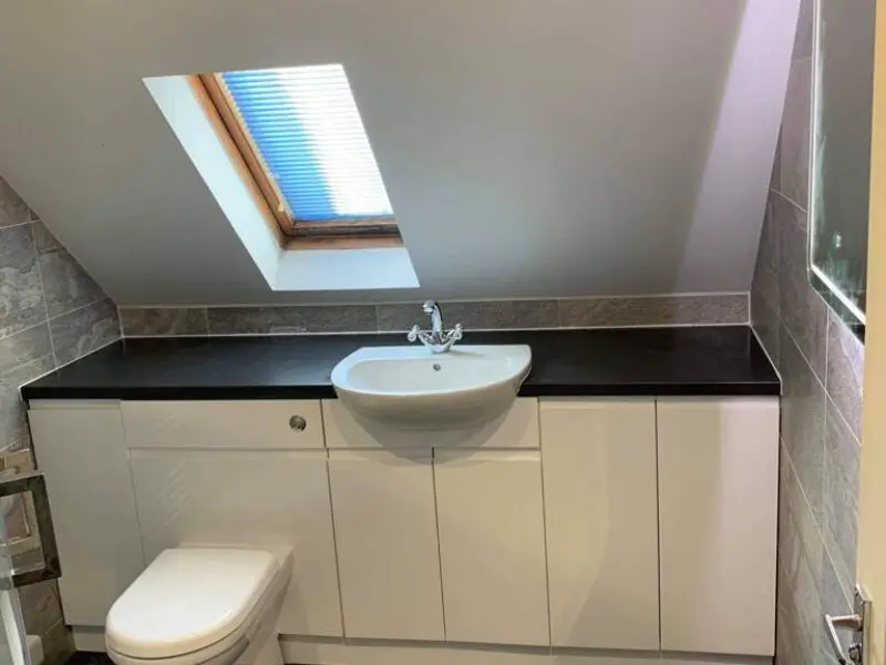 A modern bathroom with a white sink and cabinet, a white toilet, and a black countertop. A skylight window with blue blinds is on the slanted ceiling above the sink. Gray tiled walls complete the room.