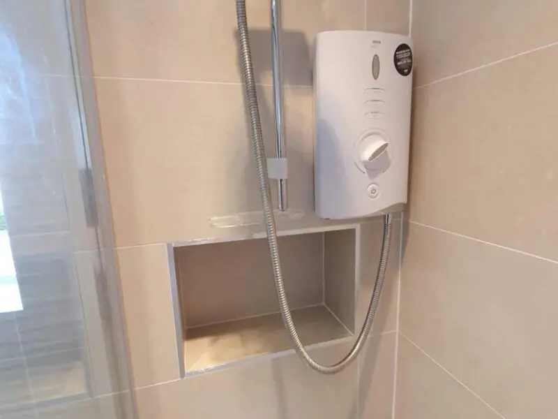 A white electric shower unit is mounted on a beige tiled wall above a recessed shelf, with the shower hose hanging down into the niche.