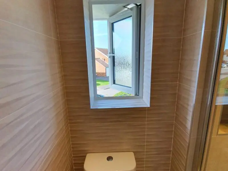 A bathroom with beige tiled walls, a white toilet below a small open window, and a view of houses and greenery outside.