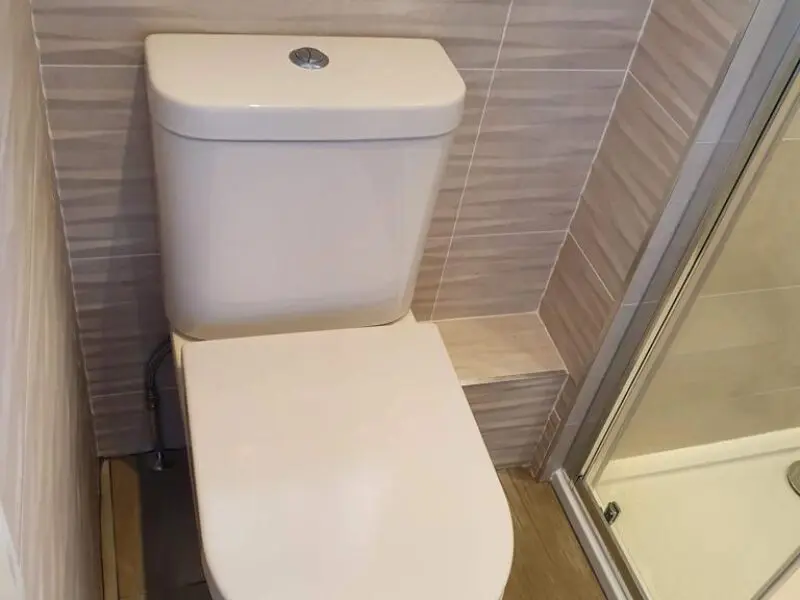 A toilet is installed in a small bathroom, with its seat partially blocked by a raised ledge on the wall, making it difficult to use. A shower enclosure with a glass door is next to the toilet.