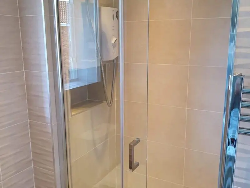 A modern shower with beige tiled walls, a glass door with a metal handle, an electric shower unit mounted on the wall, and a chrome towel rail visible on the right.