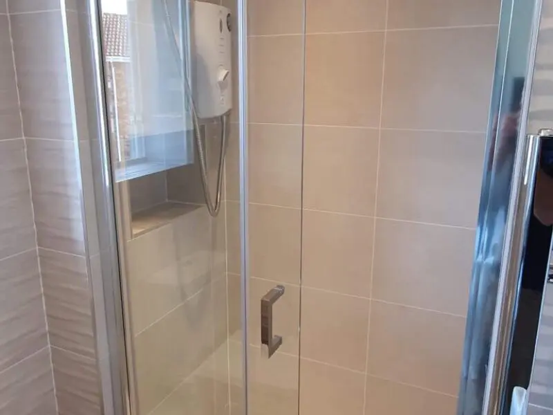 A shower with beige tiled walls, a glass door, and a mounted electric shower unit next to a small window inside the shower enclosure.