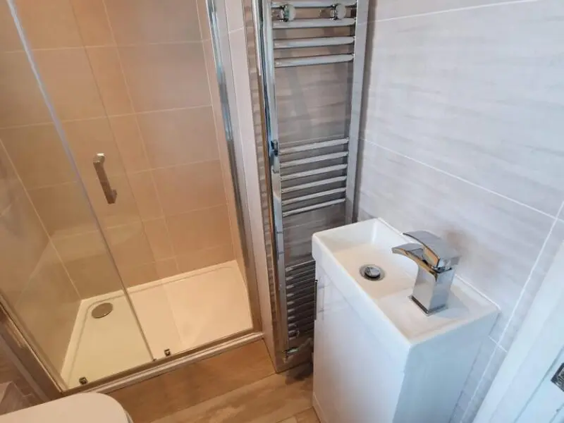 A small bathroom with a glass shower enclosure, a modern rectangular sink with a chrome faucet, and a tall, narrow heated towel rail placed between the shower and the sink.