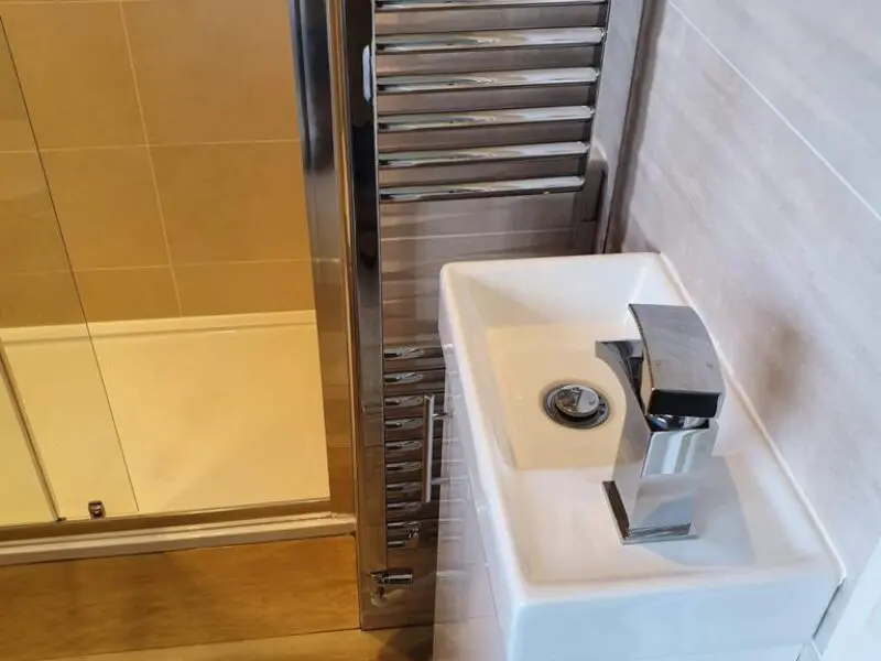 A small white sink with a modern square faucet is positioned very close to a tall chrome towel radiator in a compact bathroom. Shower doors and beige tiles are visible next to the sink.